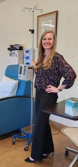 A woman with long blonde hair, wearing a patterned blouse and black pants, stands smiling next to an IV pole in a medical office. A blue chair and medical equipment are in the background.