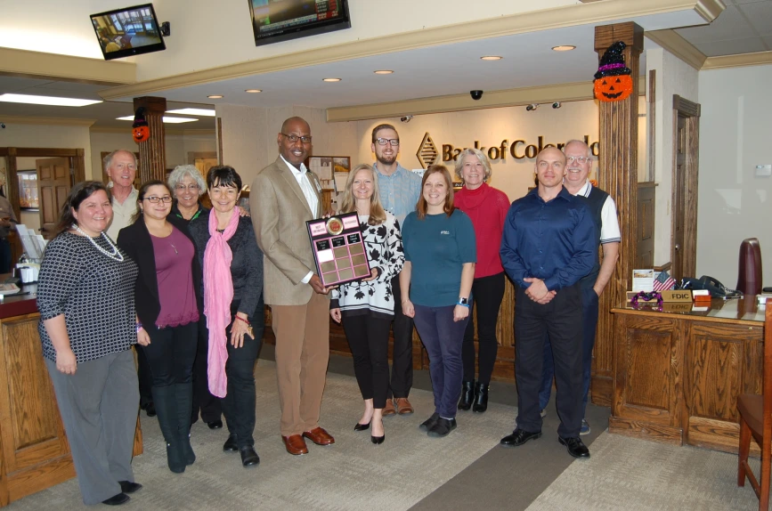 A group of people stand together indoors, holding a plaque in front of a Bank of Colorado sign with Halloween decorations around.