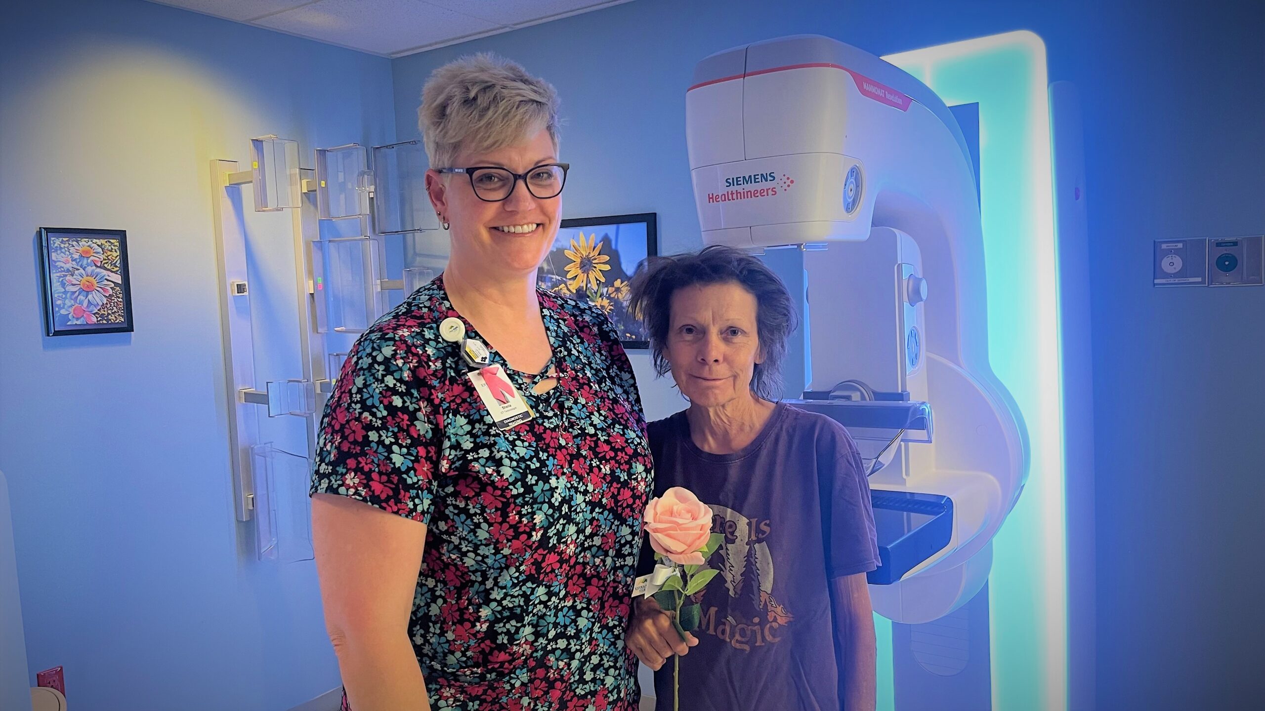 Two women stand in a medical room with a large diagnostic machine behind them. One woman holds a pink rose and the other wears medical credentials on a lanyard while smiling.