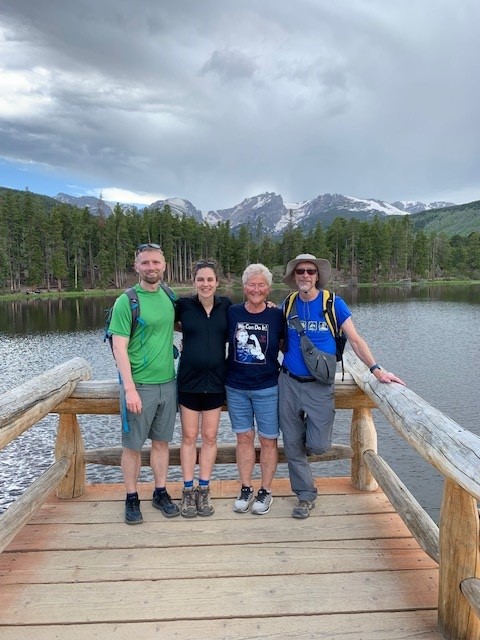 Four people stand on a wooden bridge with a lake and snow-capped mountains in the background. They are dressed in casual outdoor clothing and are smiling at the camera.