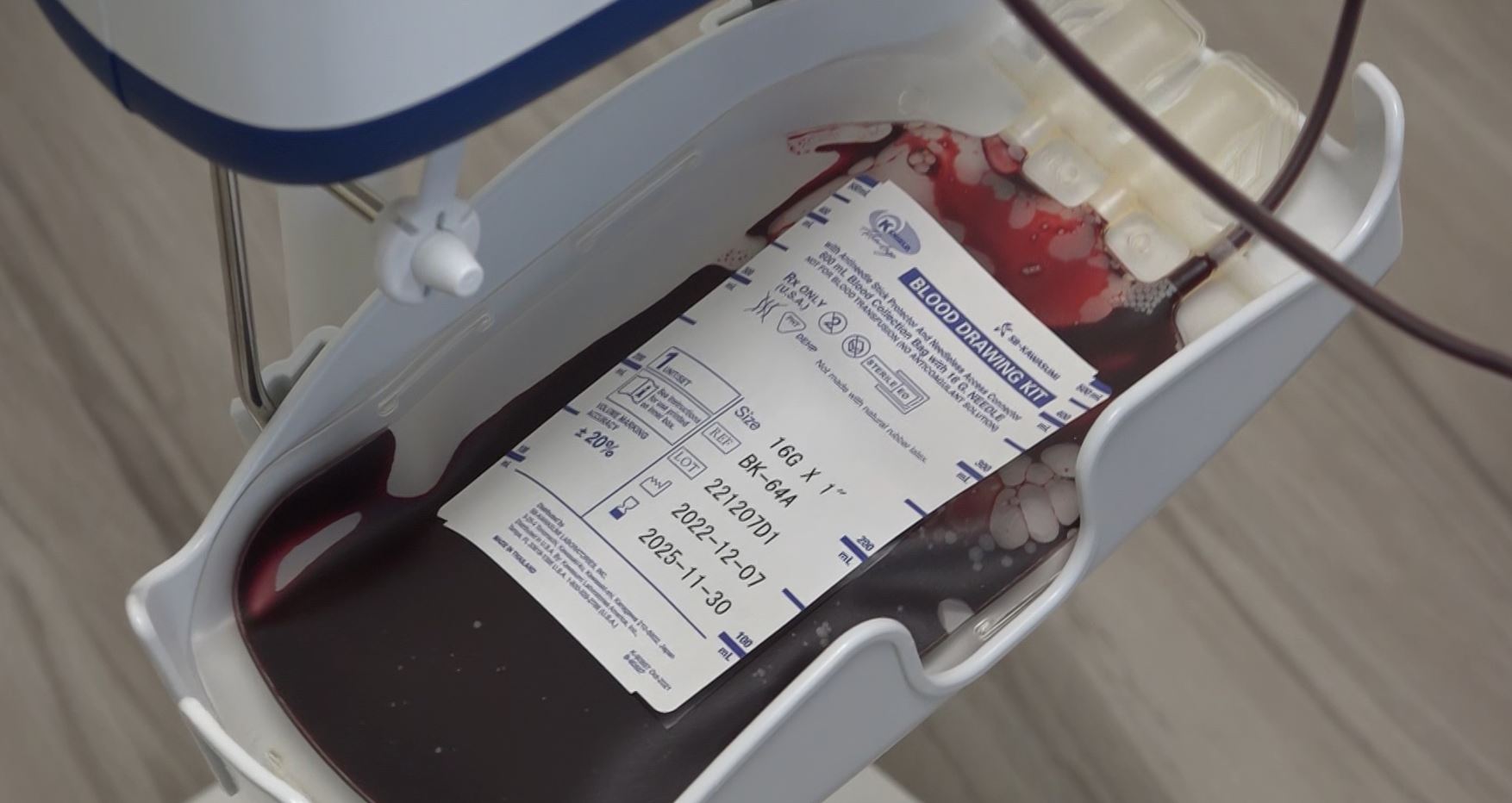 Close-up of a medical device containing a blood bag labeled with patient information, blood type, and expiration date. Blood tubes are connected to the bag, and there's blood present in the device.