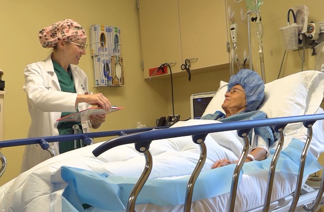 A medical professional in scrubs and a surgical cap is talking to a patient lying in a hospital bed in a clinical room filled with medical equipment.