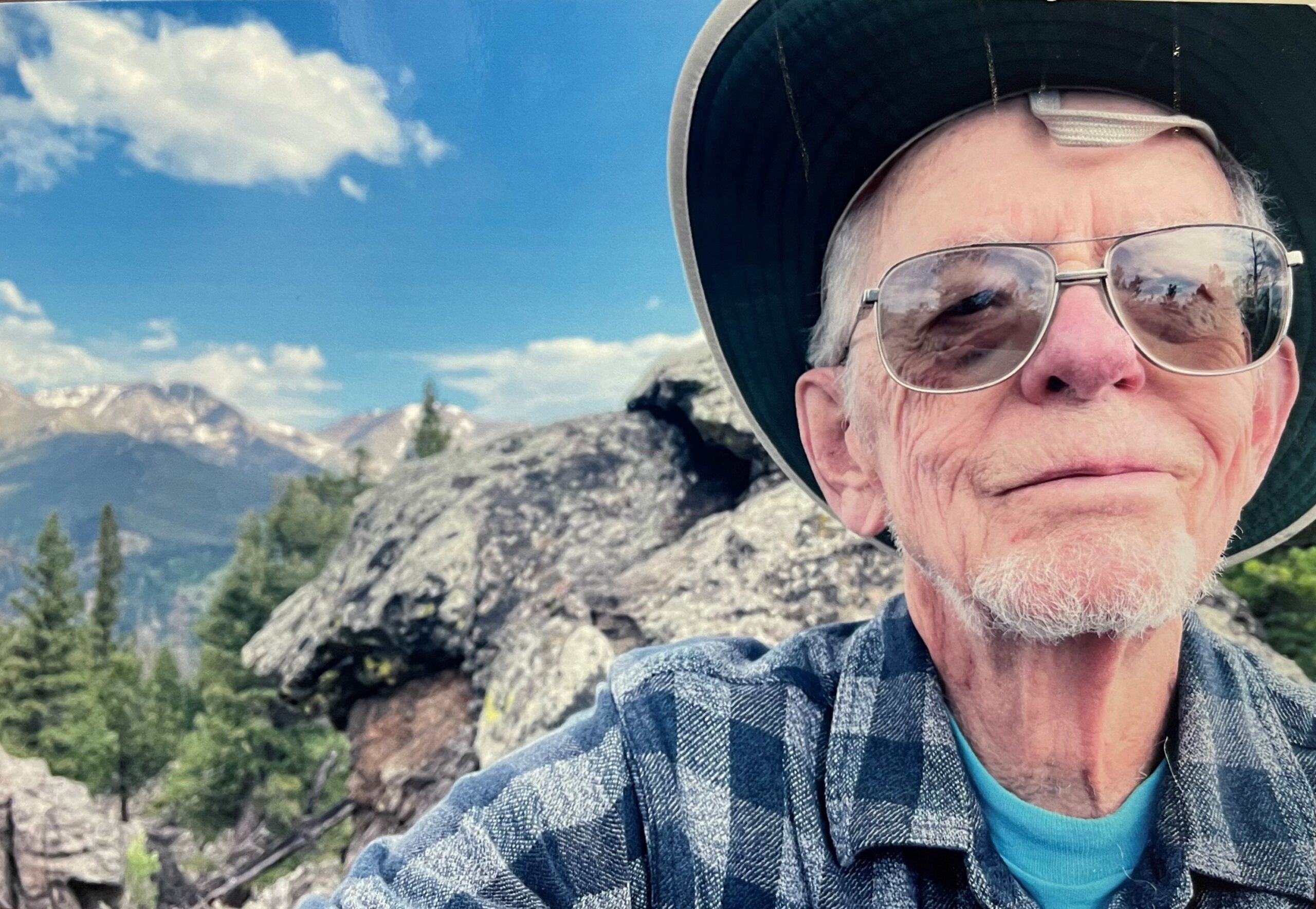 An elderly man wearing a wide-brimmed hat and sunglasses poses for a selfie in a mountainous outdoor setting with trees and rocks in the background.