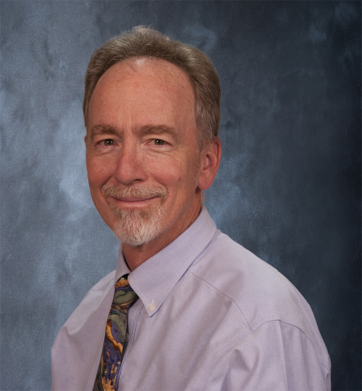 A man with gray hair and a beard, wearing a light purple shirt and a colorful tie, smiles against a dark gray textured background.