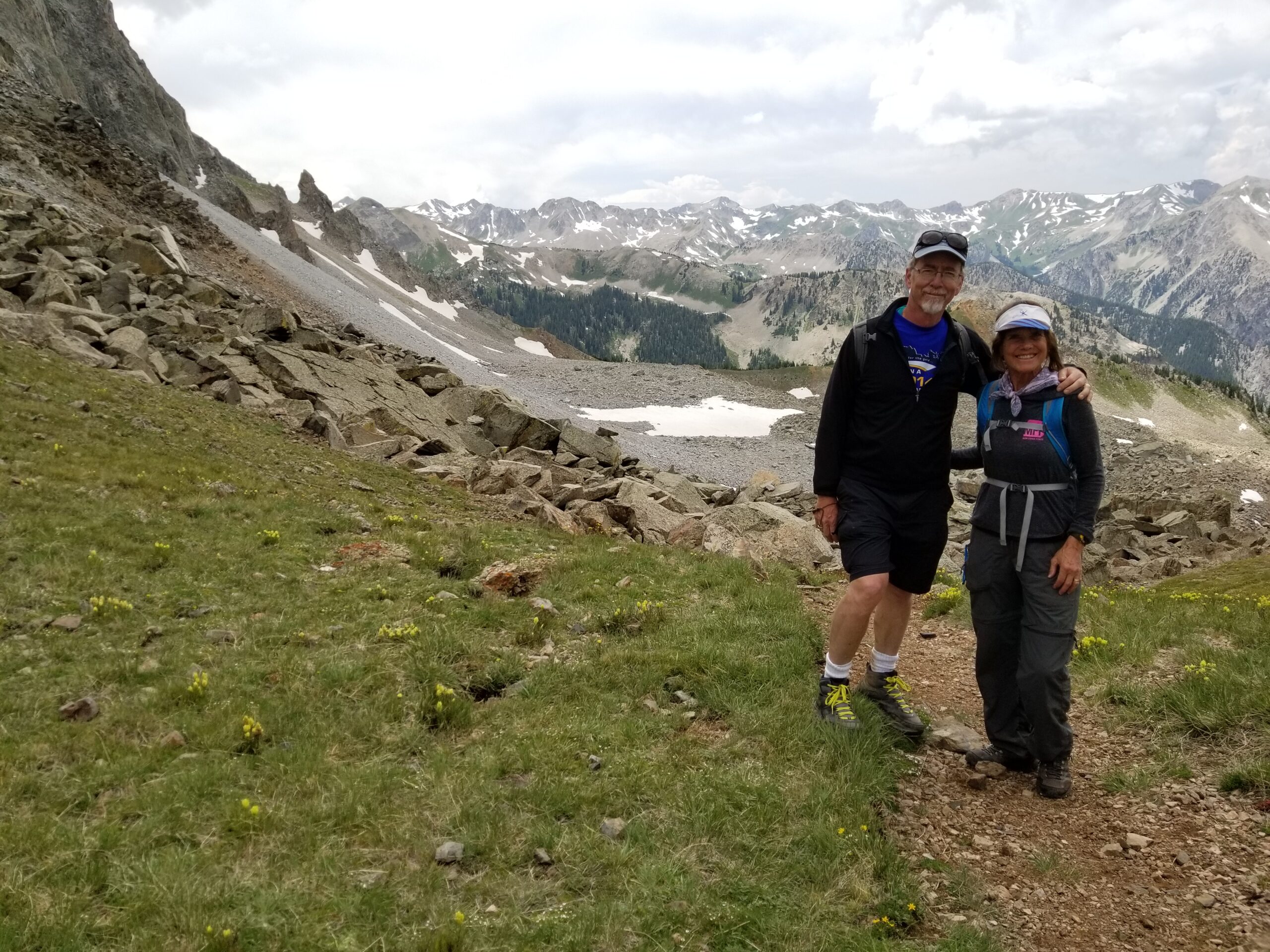 Two hikers standing on a rocky trail with mountainous terrain and scattered patches of snow in the background. Both are dressed in outdoor gear and smiling at the camera.