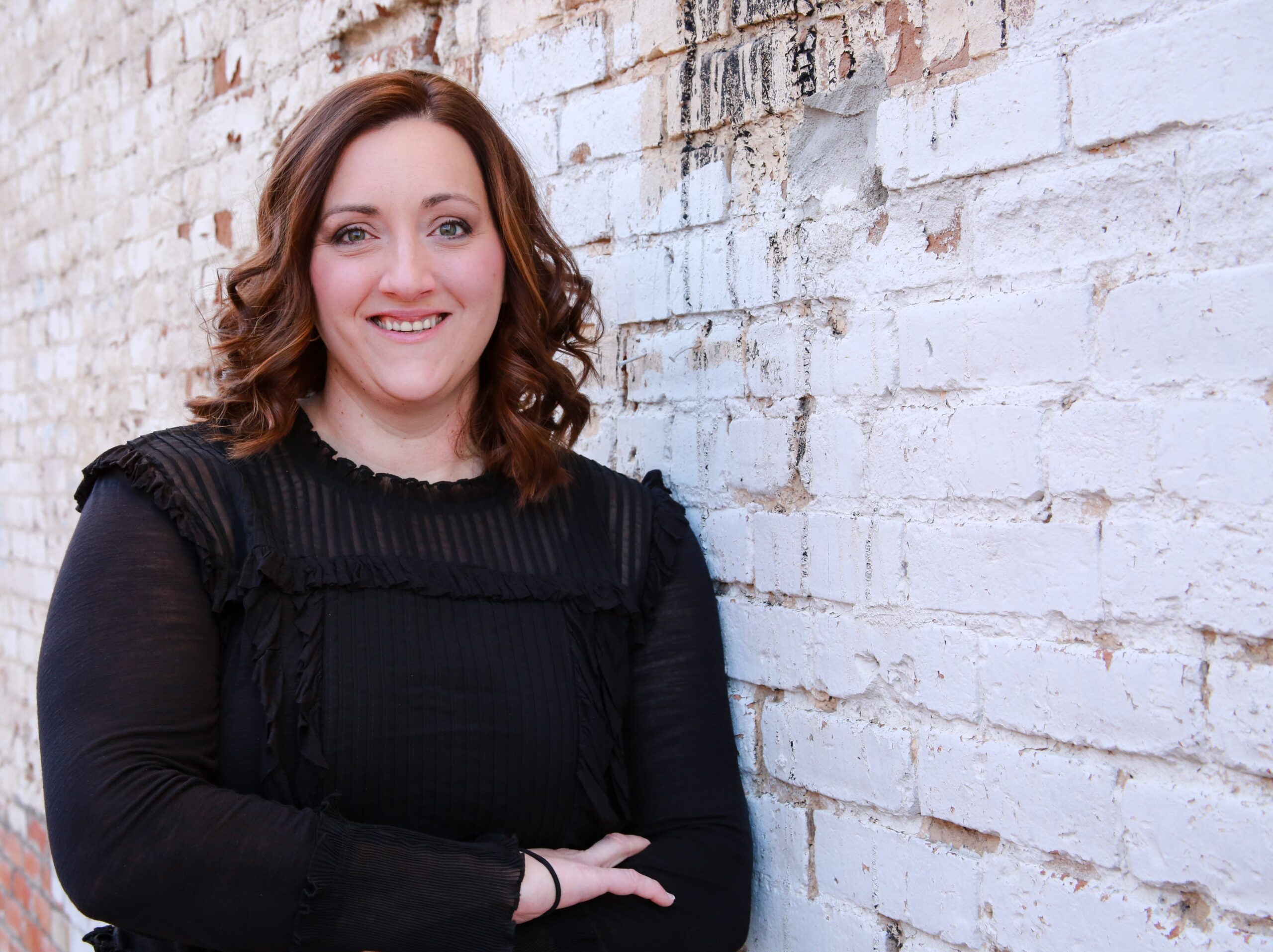 A person with shoulder-length brown hair is smiling and standing against a white brick wall, wearing a black long-sleeve top.