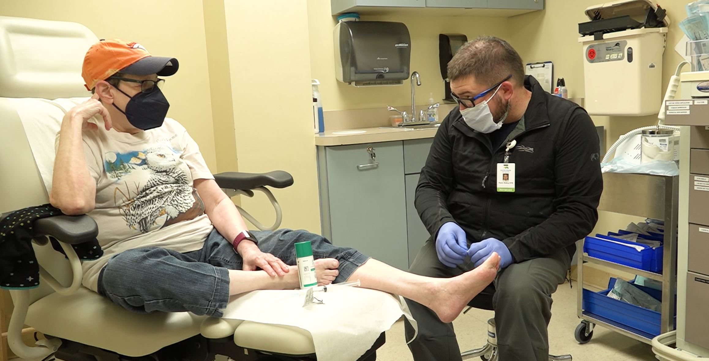 A healthcare worker in medical attire examines the foot of a seated patient in a clinical setting; both are wearing masks. The patient is in casual clothes and appears to be receiving foot care.