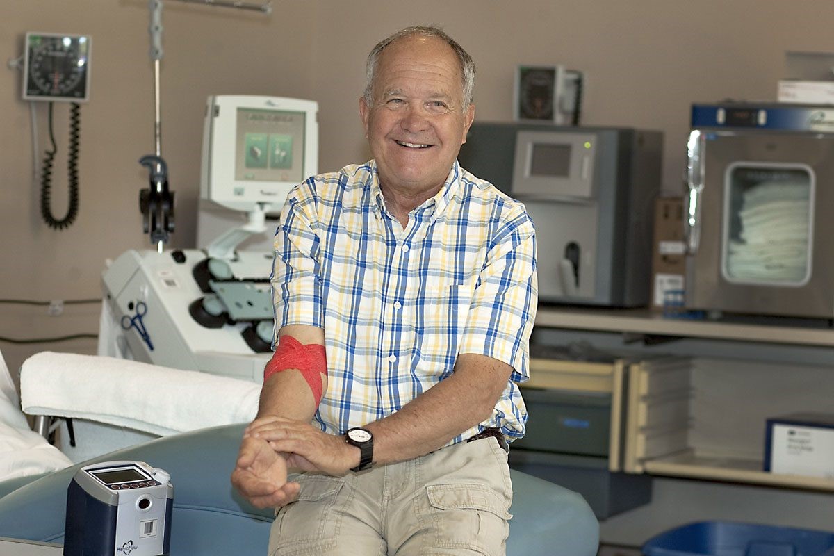 An elderly man, wearing a plaid shirt, sits in a medical office with a red bandage on his arm and smiles at the camera. Medical equipment is visible in the background.