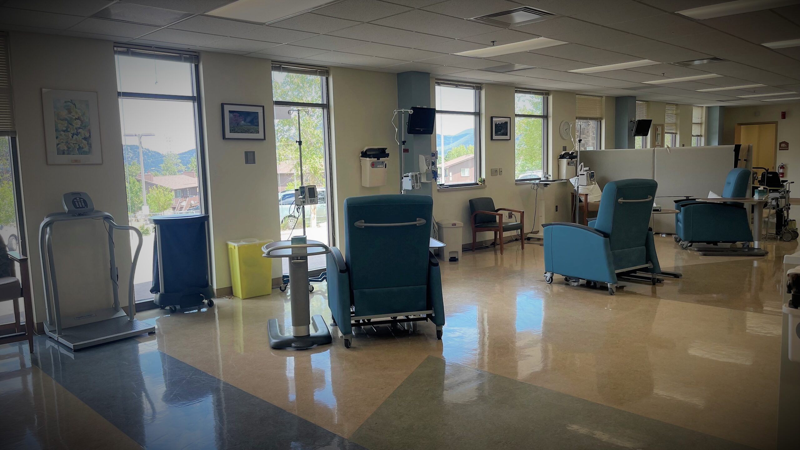 A medical clinic room with multiple recliner chairs, medical equipment, and a treadmill near large windows overlooking a scenic view. The room has polished floors and bright lighting.