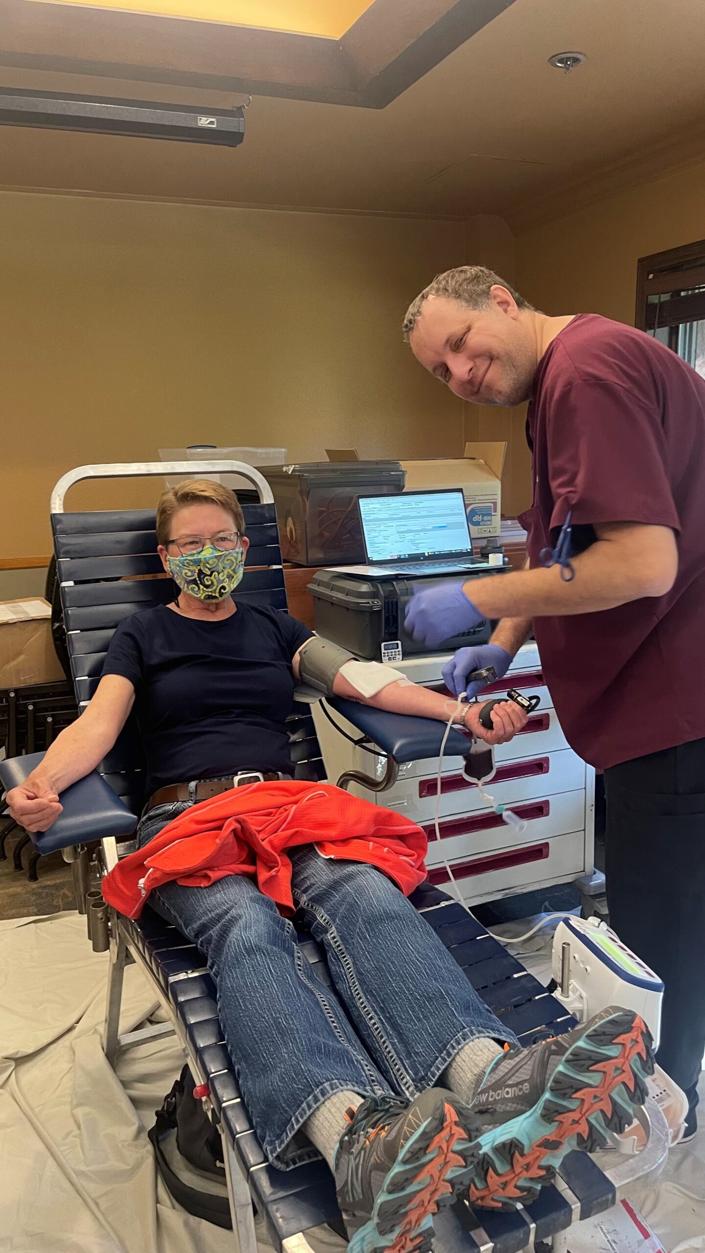 A person donates blood while a medical professional, wearing gloves, assists. The donor is seated on a reclining chair, and the medical professional is taking blood pressure readings.