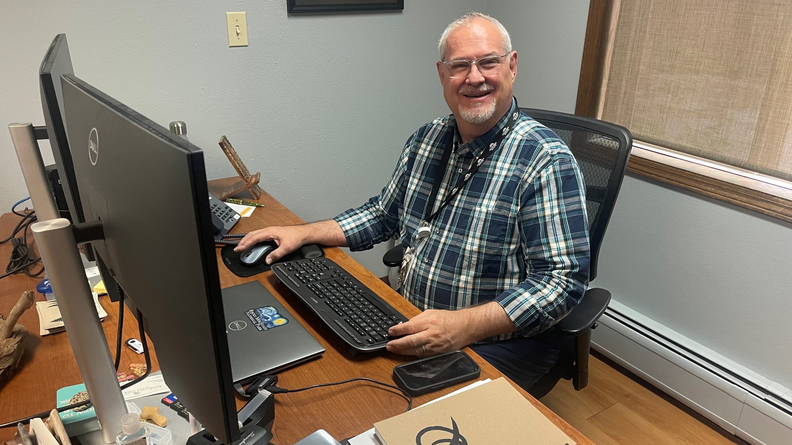 A man with glasses and a plaid shirt sits at a desk with dual monitors, a laptop, and various office supplies, smiling at the camera.