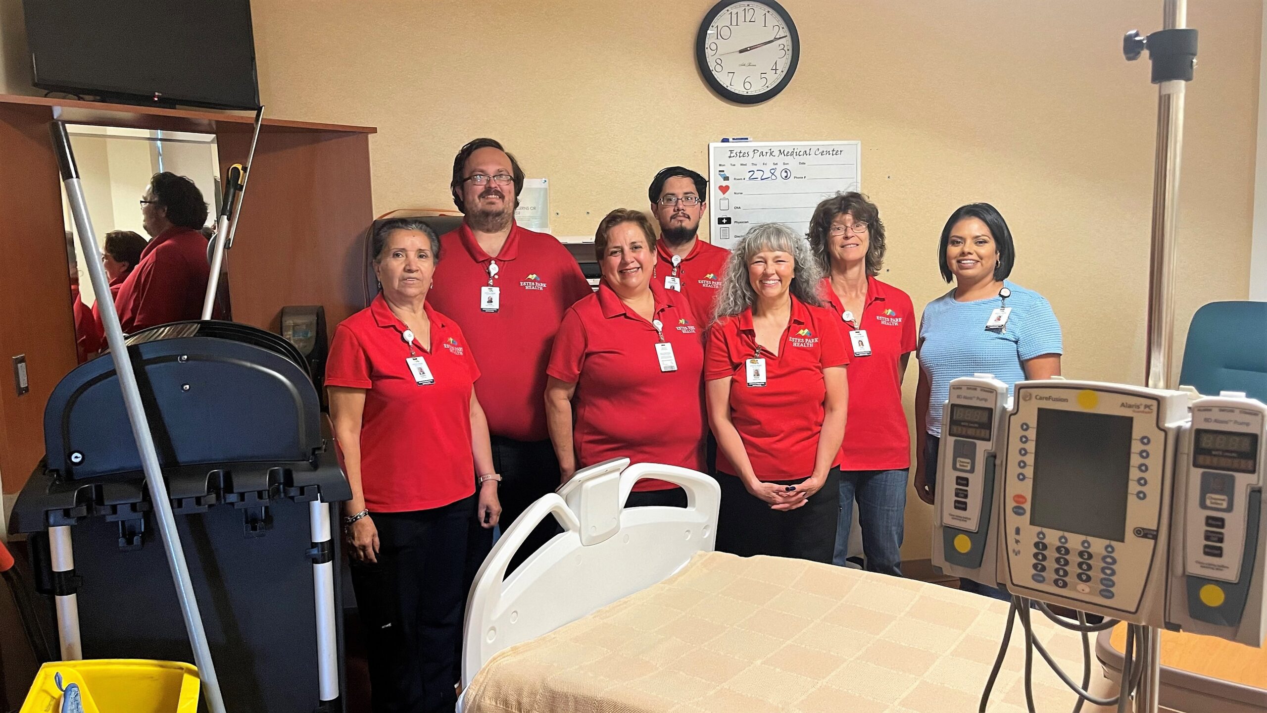 A group of seven hospital staff, six wearing red shirts and name badges, and one in blue, standing in a hospital room with medical equipment and a bed. A clock and a whiteboard are visible in the background.