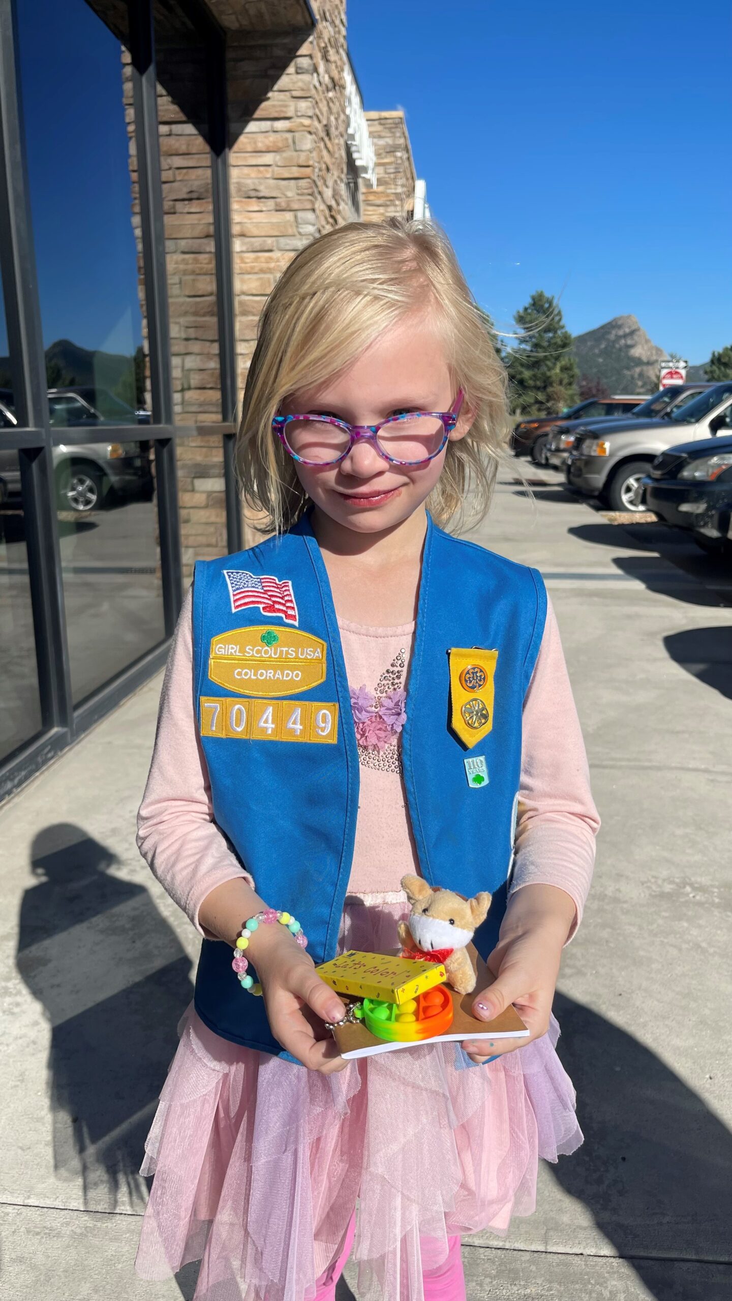 A young girl wearing glasses and a Girl Scouts vest stands outside a building on a sunny day, holding a small toy. A parking lot and a mountain are visible in the background.