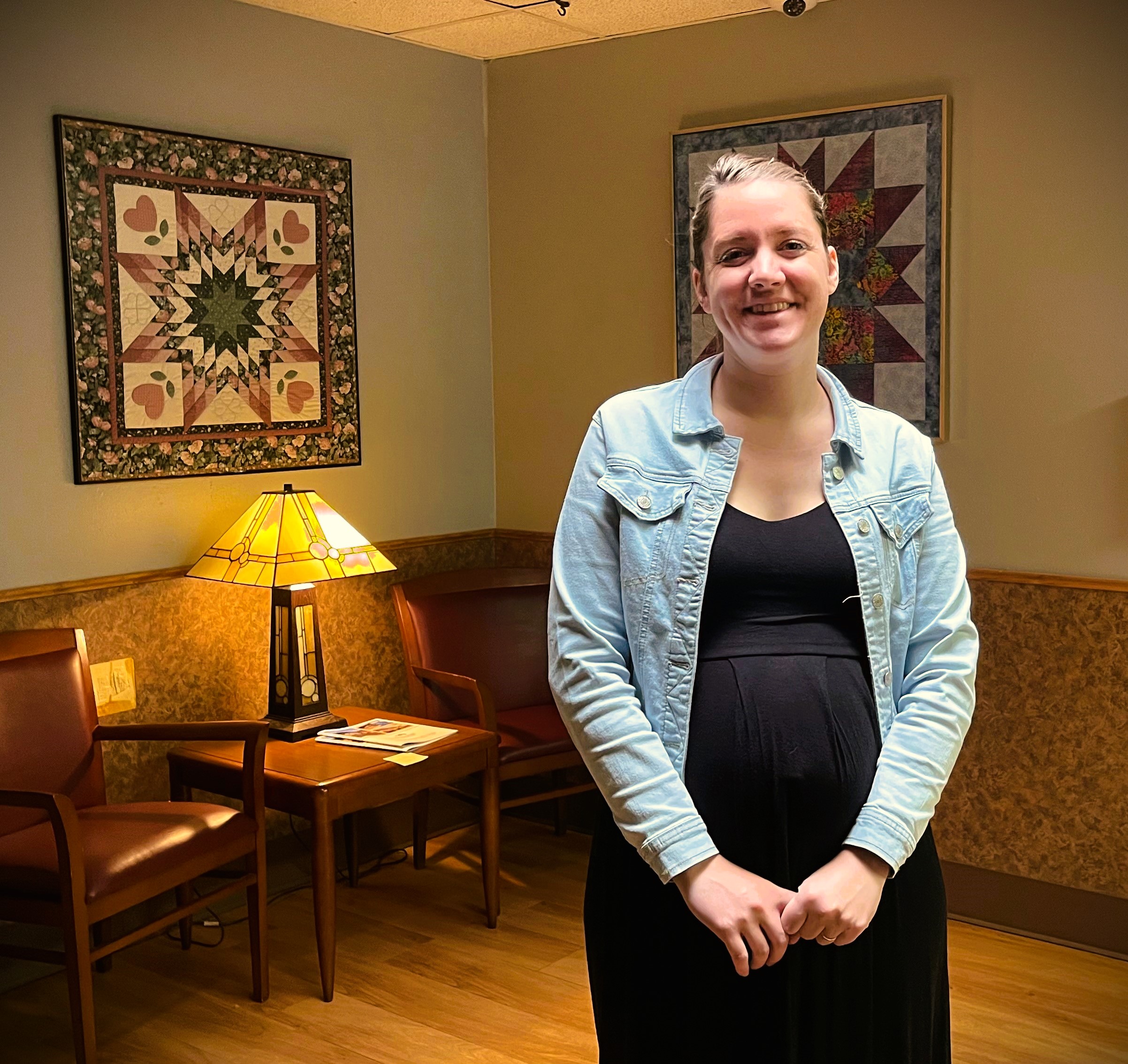 A person stands in a waiting room with quilted wall hangings, a table with a lamp and magazines, and wooden chairs. They are smiling and wearing a denim jacket over a black dress.