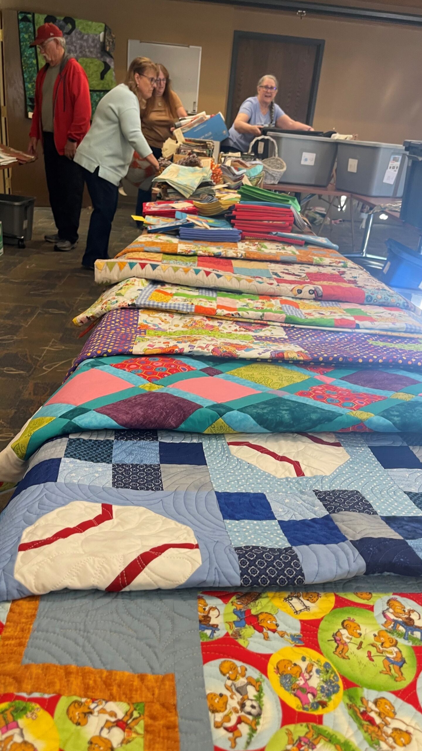 People sorting through items on a long table covered with colorful quilts, with bins and other materials around. The setting appears to be indoors.