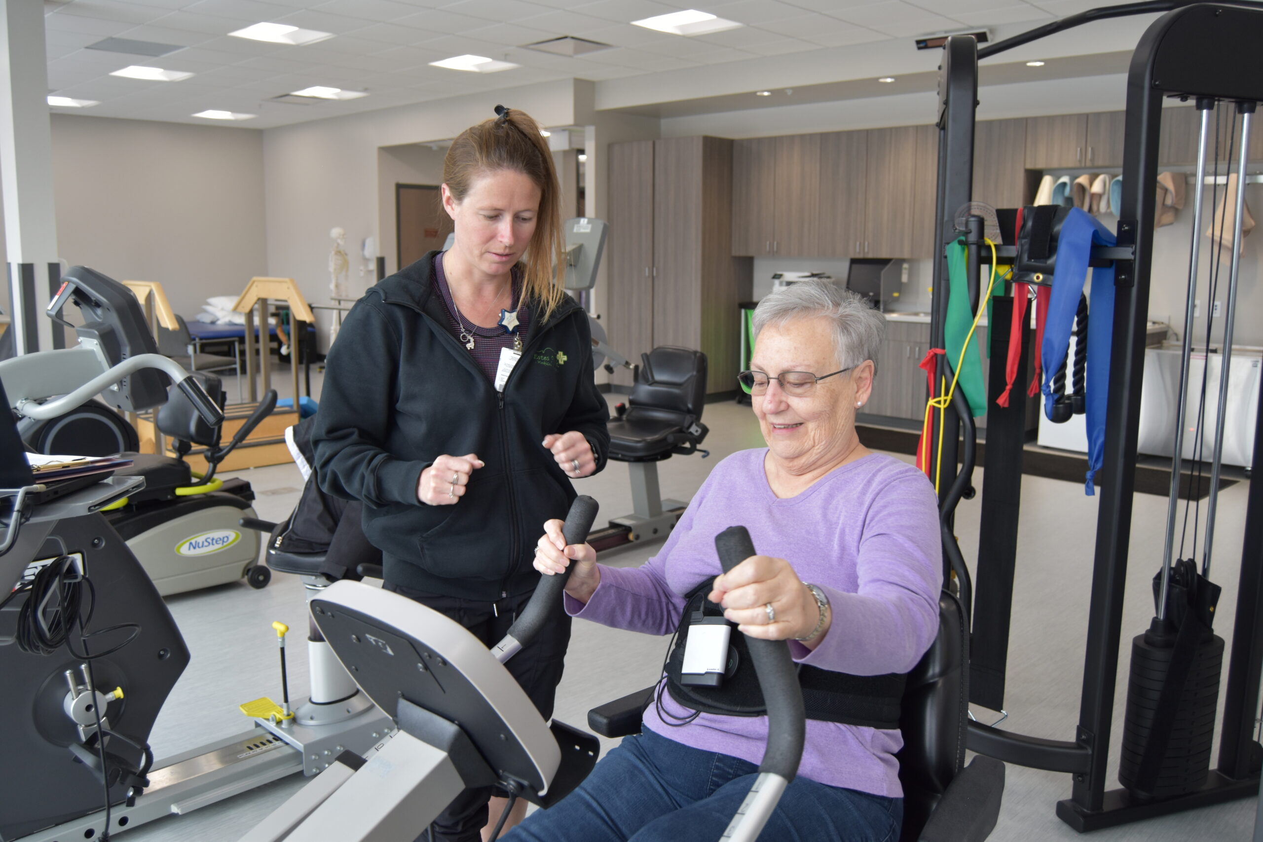 An elderly woman exercises on a seated machine while being assisted by a trainer in a gym. The gym has various exercise equipment.