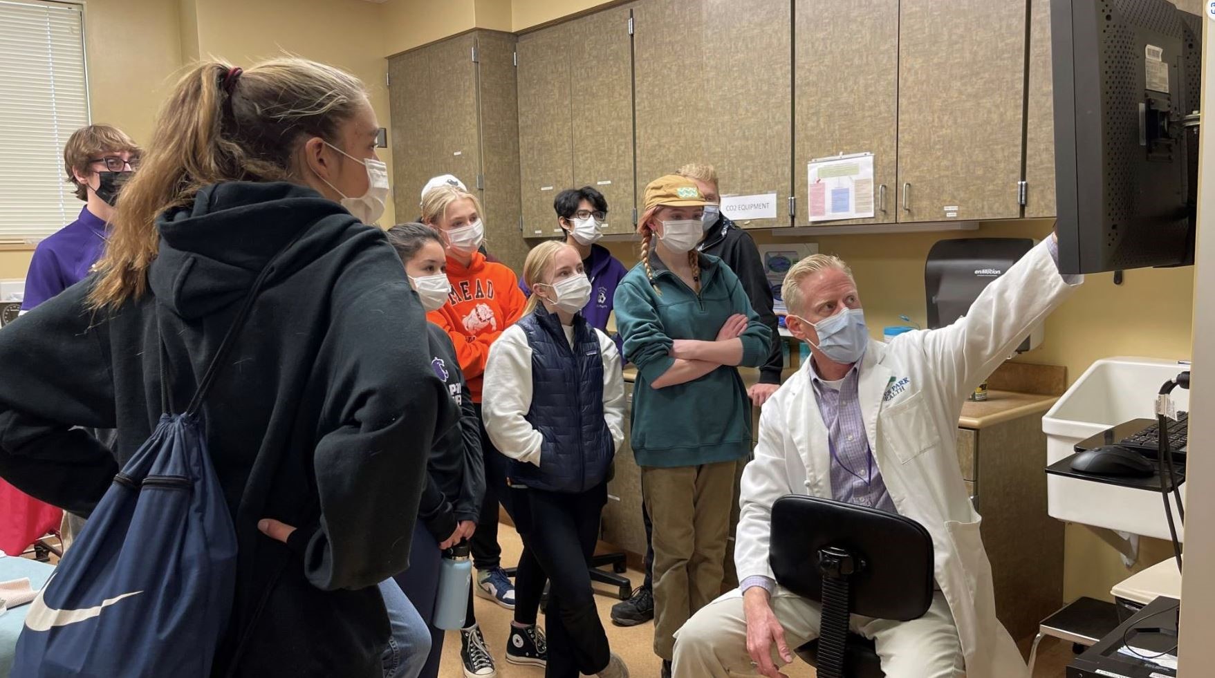 A group of people in masks listens to a man in a white coat who is pointing at a computer screen in an office setting.