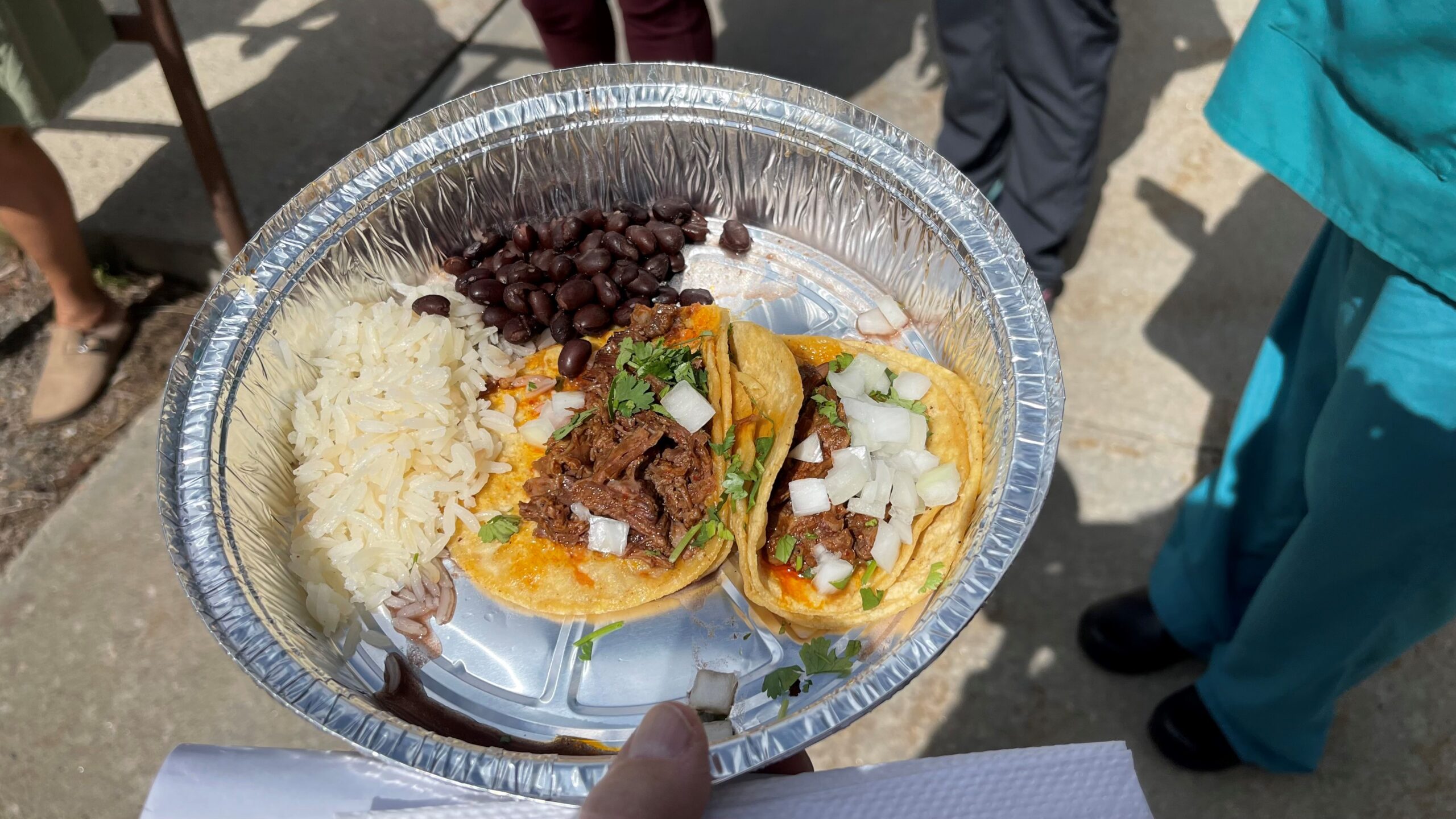 A foil container with two tacos topped with diced onions and cilantro, served with white rice and black beans.