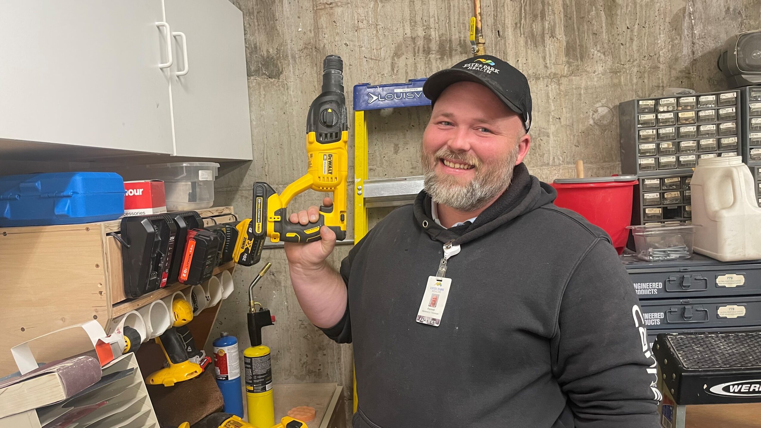 A man wearing a black hoodie and cap smiles while holding a yellow cordless drill in a workshop setting. Various tools and equipment are visible in the background.