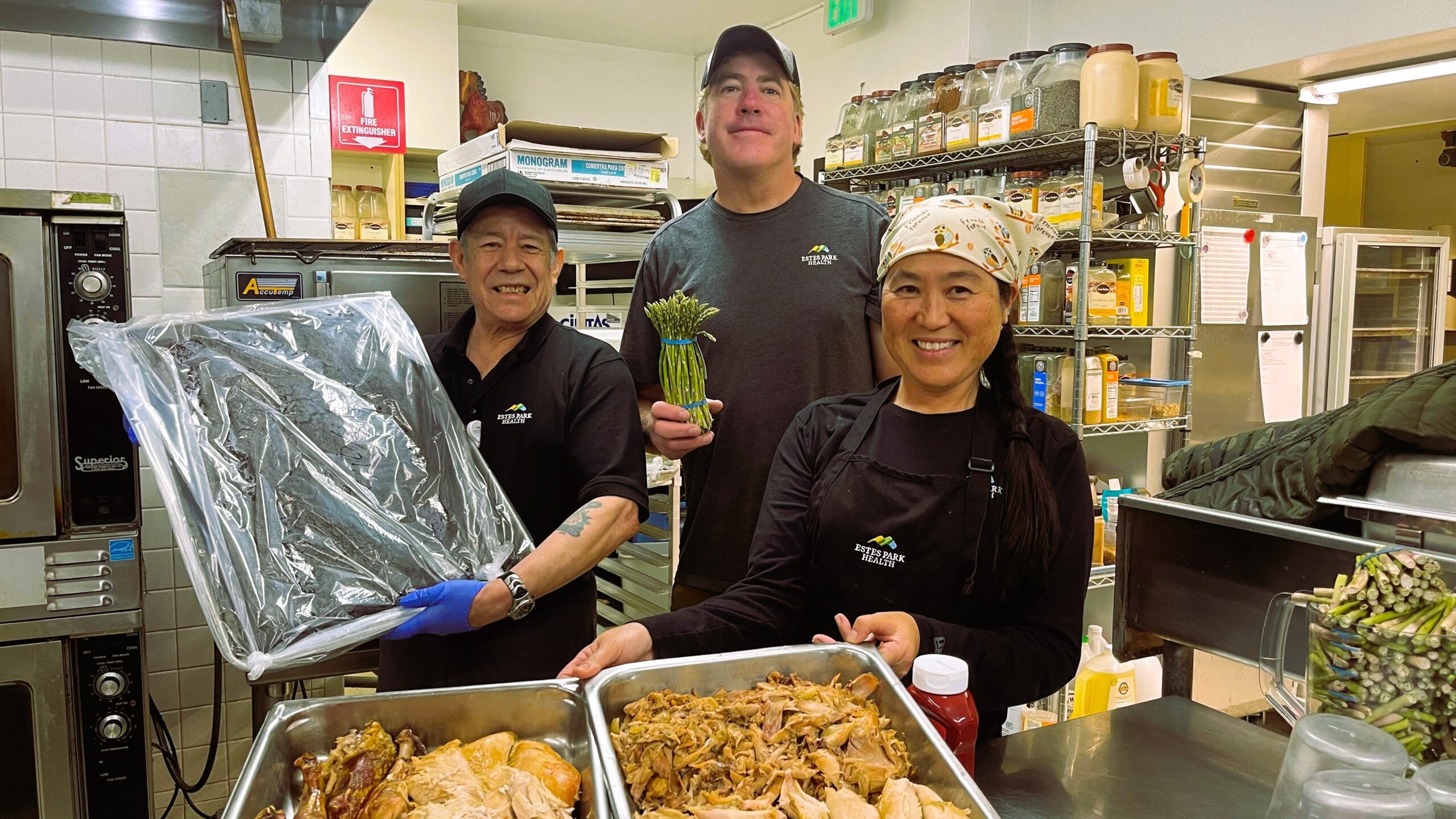 Three kitchen staff members in aprons, smiling at the camera, holding trays of prepared food. One holds asparagus, another holds a tray covered in foil, and the third a tray with roast poultry.