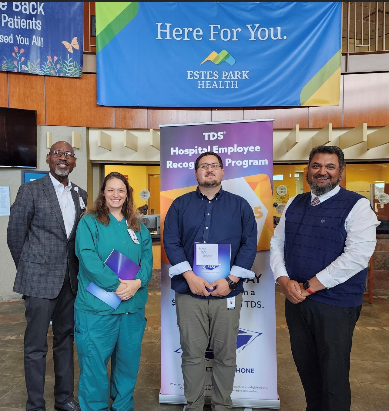 Four people pose for a photo in front of an Estes Park Health banner and a TDS Hospital Employee Recognition Program sign. One person is in medical scrubs, others in business attire, all smiling.