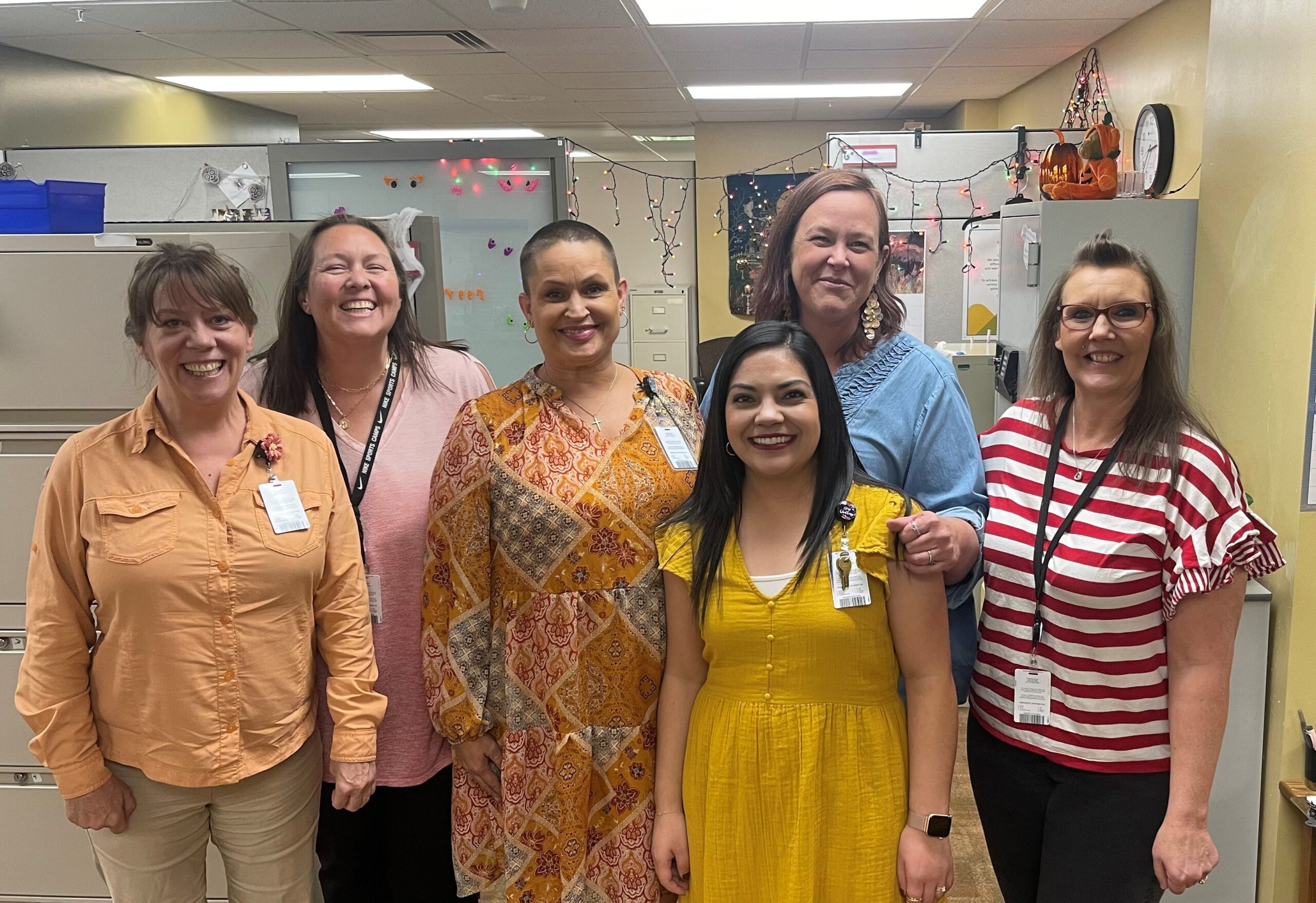 Five women and one man standing in an office wearing varied outfits, each with a name tag, smiling at the camera. The office is decorated with Halloween-themed decorations.