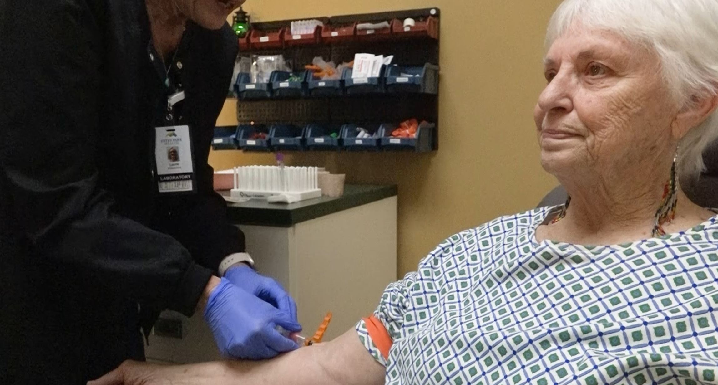 A healthcare professional prepares to draw blood from an elderly woman seated in a medical clinic. Equipment and supplies are visible in the background.