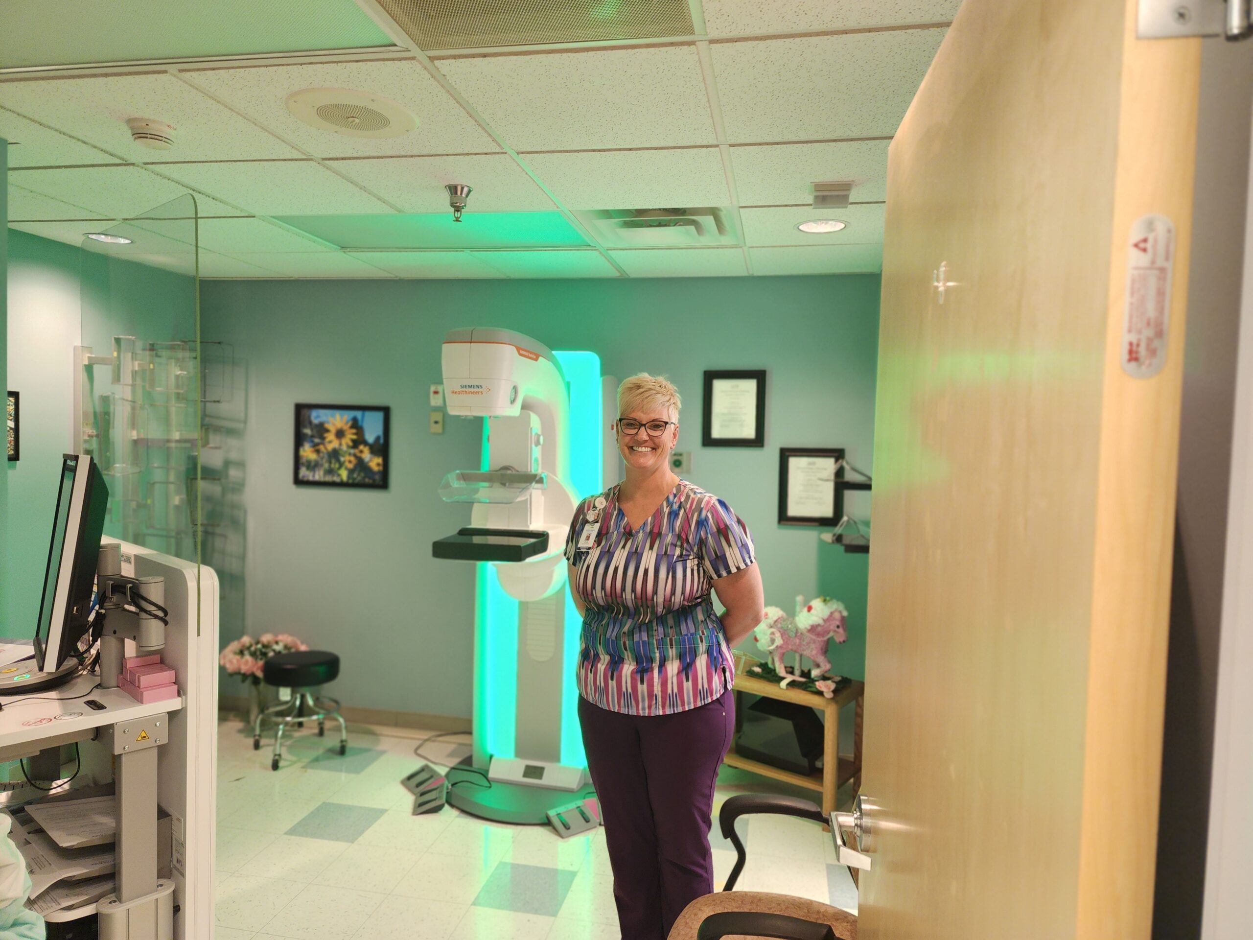A woman wearing a colorful top and glasses stands in a room with medical equipment, including a mammography machine. She smiles, positioned next to a partially open door.