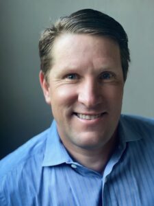 A man with short brown hair and blue eyes is smiling. He is wearing a light blue button-up shirt.