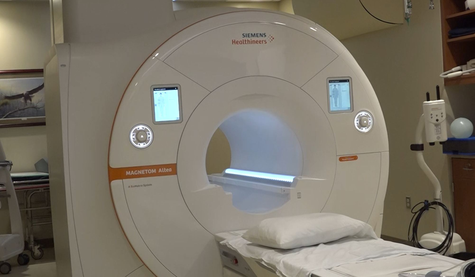 A Siemens Healthineers MAGNETOM Altea MRI machine in a hospital room with a pillow on the patient bed and various medical equipment around.