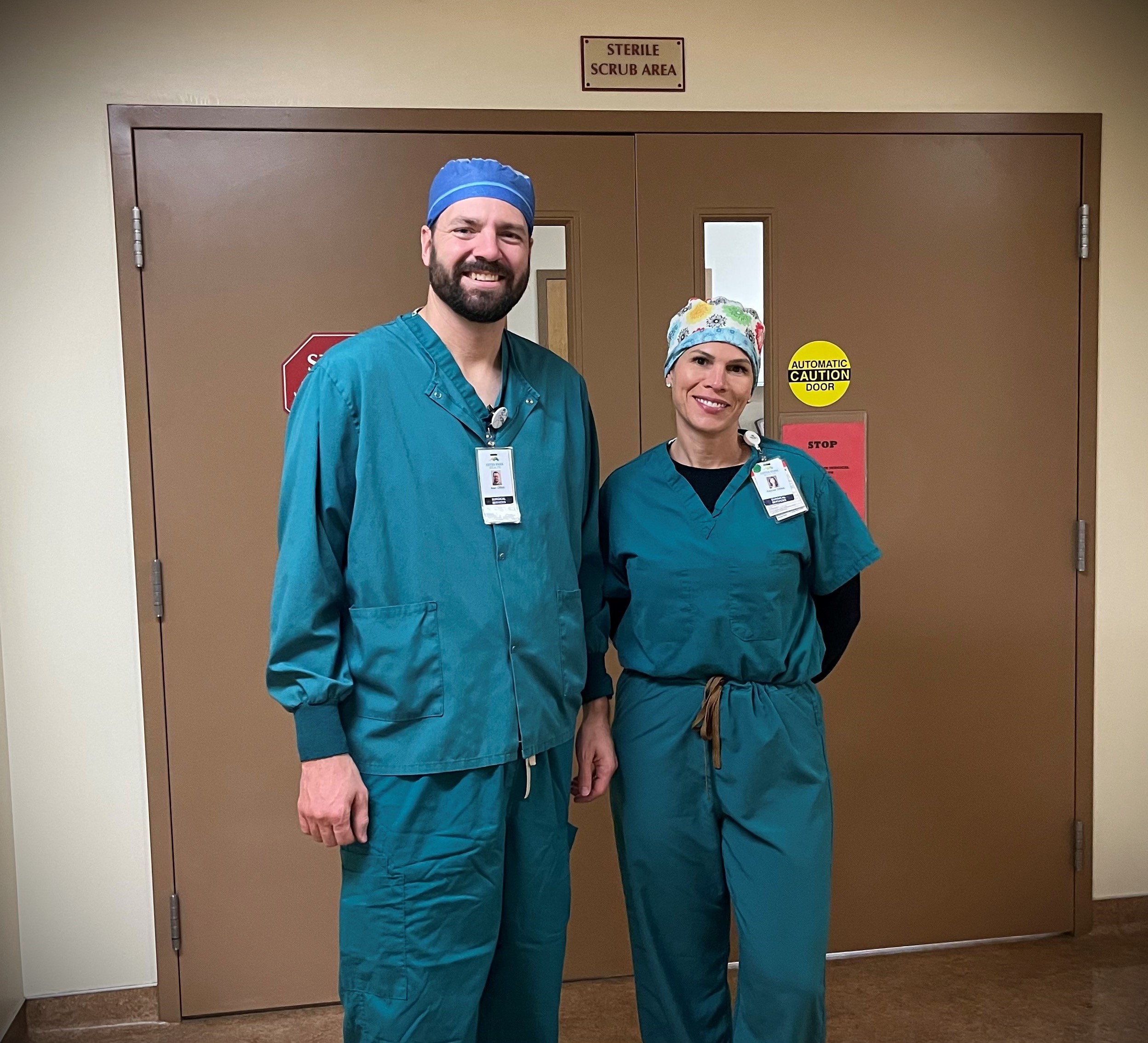 Two medical professionals in green scrubs and surgical caps stand in front of doors marked 