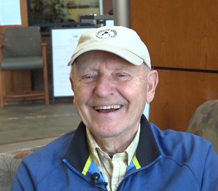 An older man wearing a beige cap and blue jacket smiles while sitting indoors. A board and chair are visible in the background.