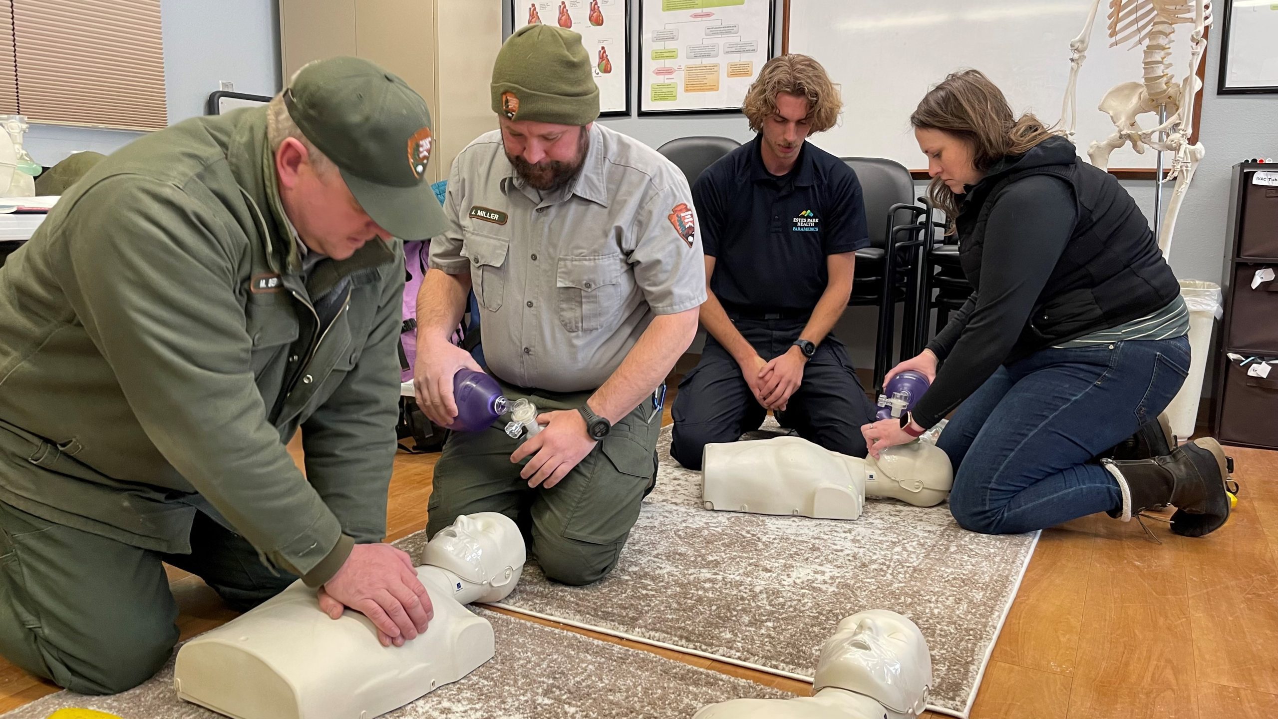 Four people practice CPR on mannequins in a training room. Two individuals use bag valve masks while the others prepare for chest compressions. They are dressed in uniforms and casual attire.