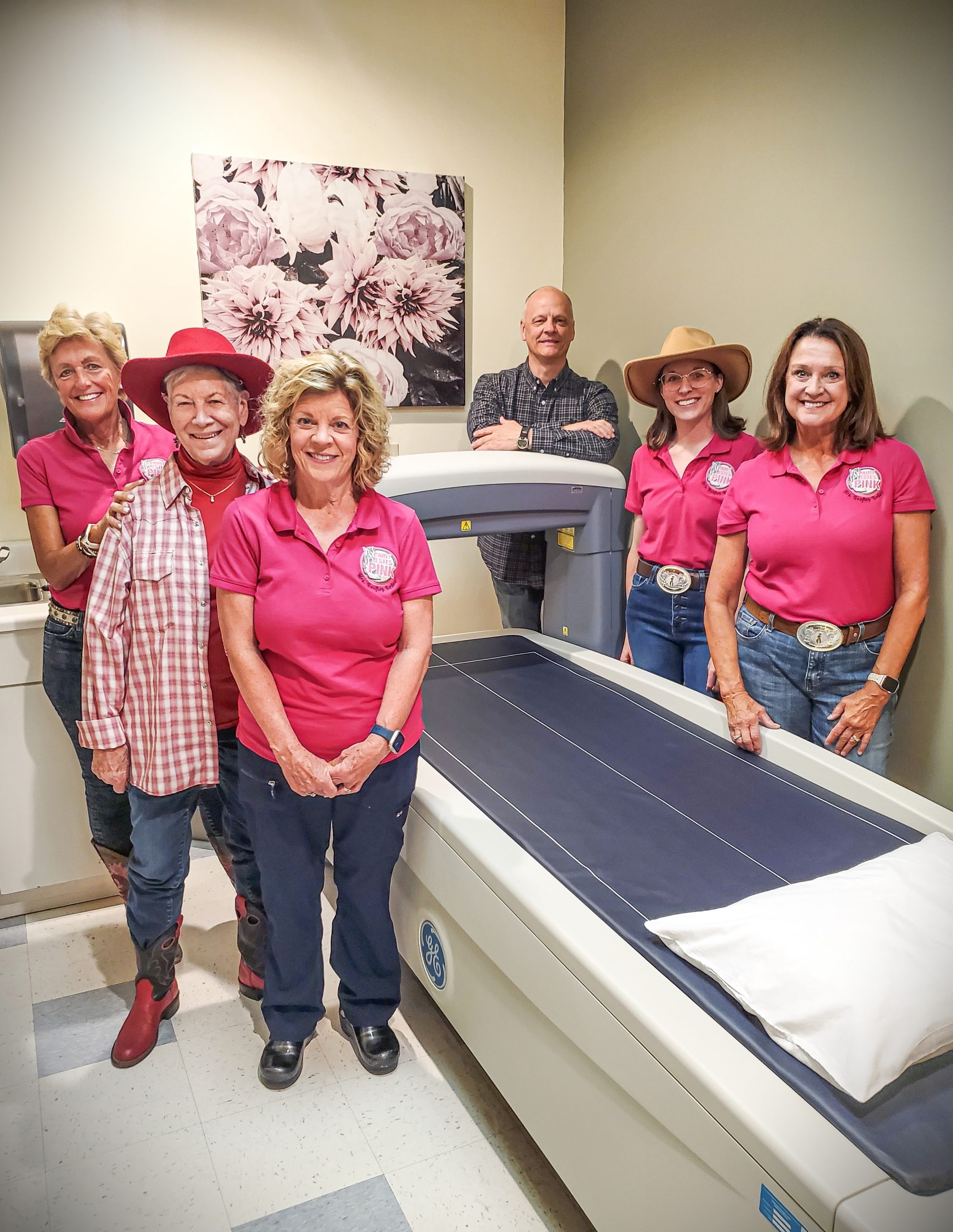 A group of five women and one man stand next to a medical machine in a room with a floral painting on the wall. The women are wearing matching pink shirts and cowboy hats.