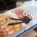 A close-up of two open boxes of assorted donuts displayed on a table, with black tongs resting in one of the boxes.