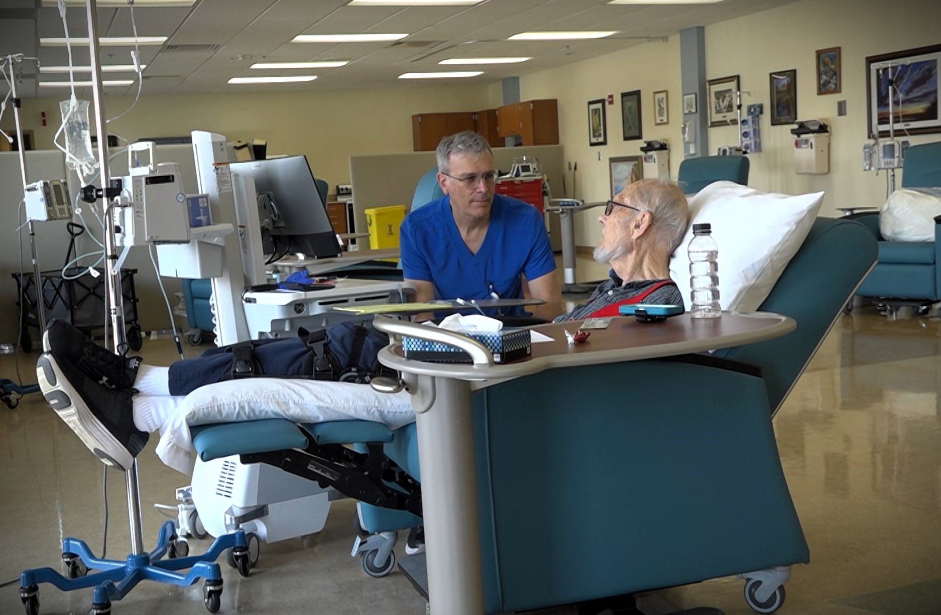 An elderly man lies on a medical recliner receiving treatment while conversing with a healthcare professional in a treatment room equipped with medical devices.