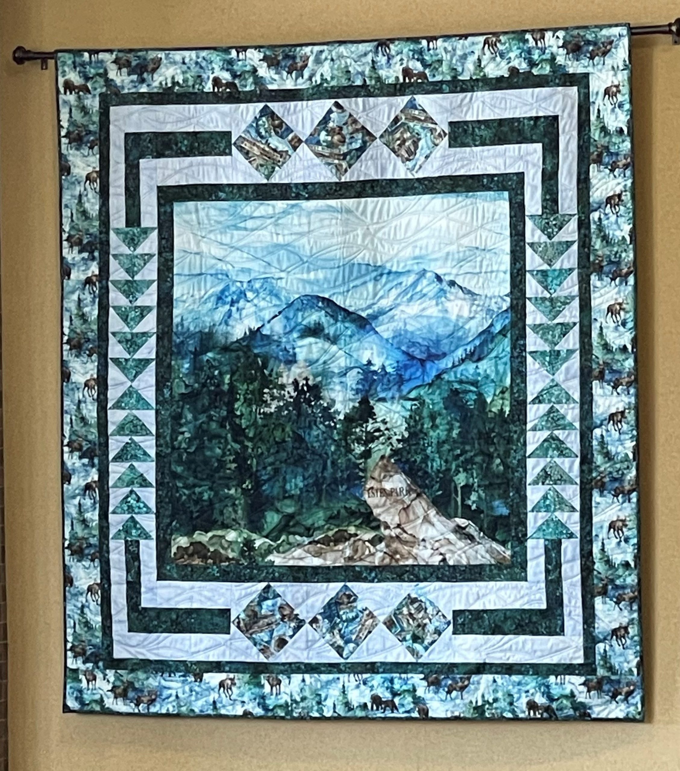 A wall-hanging quilt featuring a central scenic mountain and forest motif, surrounded by geometric border patterns in shades of blue, green, and white.