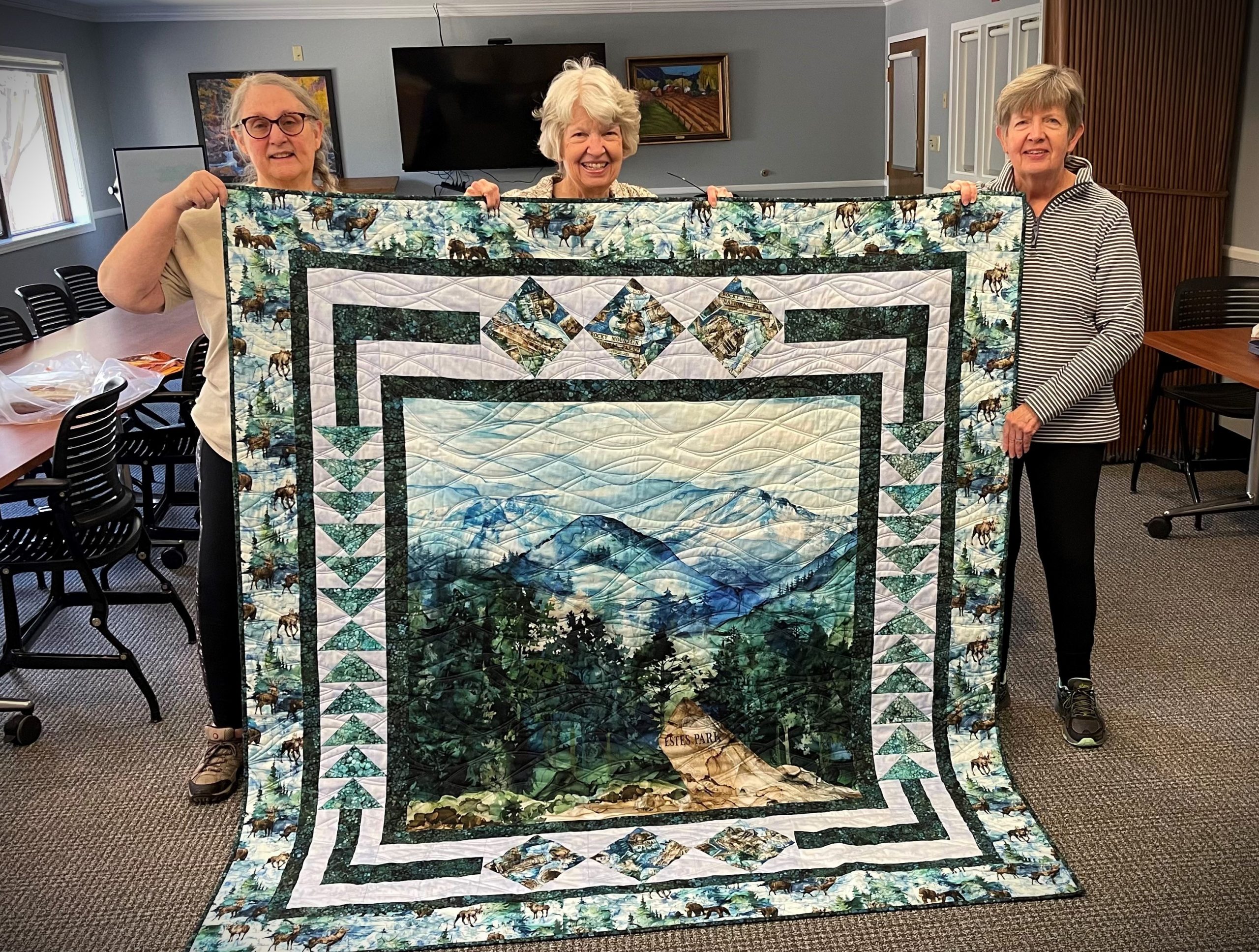 Three women hold up a large quilt featuring a mountain landscape with a trail. The setting appears to be a room with tables and chairs.