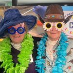 Two people wearing humorous hats, large novelty glasses, and colorful leis while standing in front of a healthcare-themed background.