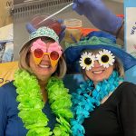 Two people wearing colorful leis, decorative sunglasses, and hats pose in front of a healthcare-themed backdrop.