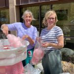 Two women stand outside by a cotton candy machine, smiling while making and bagging pink cotton candy. They are in front of a building with large windows reflecting the scene.