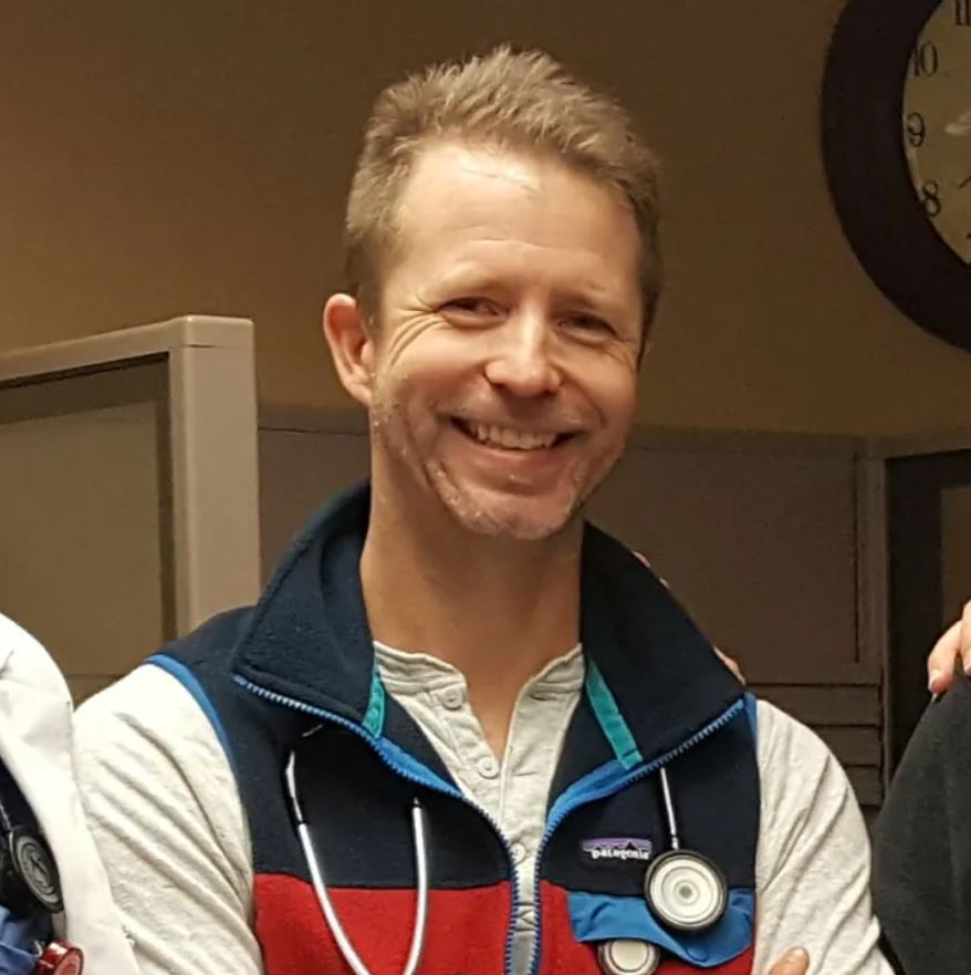 A person with short hair is smiling, wearing a jacket with a stethoscope around their neck. A clock is visible in the background.