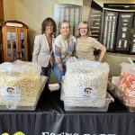 Three women stand behind a table featuring large bins of popcorn at a promotional event. Two signs on the wall behind them list names.