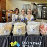 Three people wearing gloves stand behind a table with large bins of popcorn at an event by Estes Park Health. Two are holding cups of popcorn while smiling at the camera.