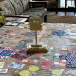 A table covered in colorful drawings and notes, with a puzzle piece trophy labeled "Puzzle Design Winner" in the center. In the background, there is a patterned couch and a large window.
