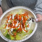 A person holding a foil bowl containing a salad with lettuce, dressing, and a red sauce.