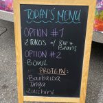 A chalkboard menu stands in front of a colorful food truck, listing two options: "#1 - 2 tacos with rice and beans" and "#2 - bowl." Protein options include barbacoa, tinga, and zucchini.