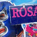 A vibrant mural features a masked luchador's face next to a purple sign with "Rosa" in pink letters.