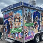 A colorful food truck, Rosa Tacos, displays vibrant artwork featuring Mexican cultural elements. People are standing nearby waiting to order. Social media icons are visible on the truck's side.