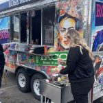 Two women at a colorful taco truck called Rosa Tacos. One woman is placing an order at the window while the other prepares food at a small outdoor station attached to the truck.